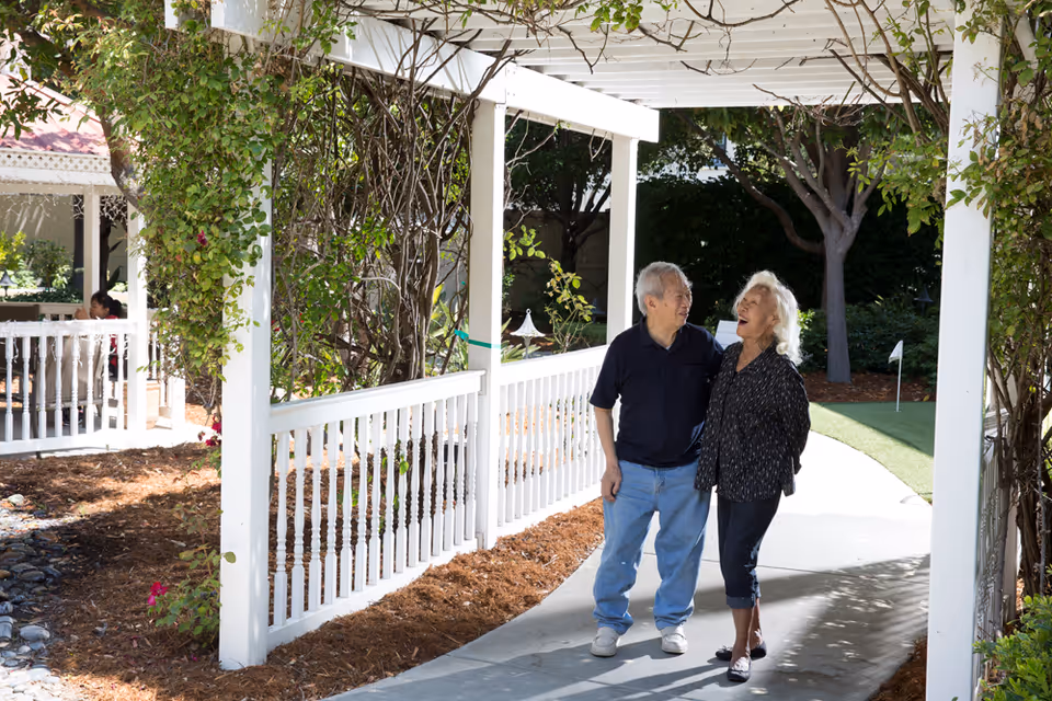 An elderly couple walking and smiling under a white pergola with climbing plants in a garden area. There are trees and a putting green in the background, and a person sitting in a gazebo to the left.