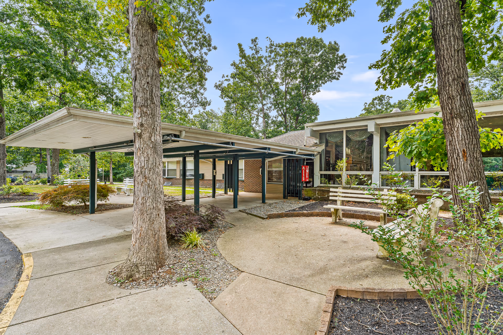 Outdoor view of a senior living facility showing a covered walkway supported by metal posts, surrounded by trees and landscaped areas with bushes and plants. There is a concrete path leading to the entrance of a building with large windows and a bench nearby.