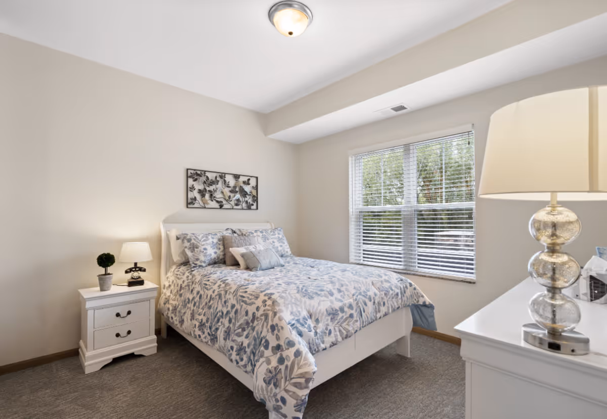 Bright, well-lit bedroom with a floral-patterned bed, nightstand, dresser and a window with blinds.