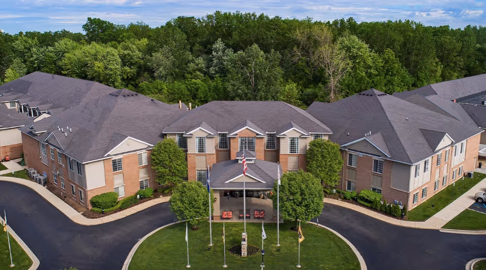Aerial front view of a two-story brick senior living facility with a circular driveway, flagpoles, and surrounding trees.
