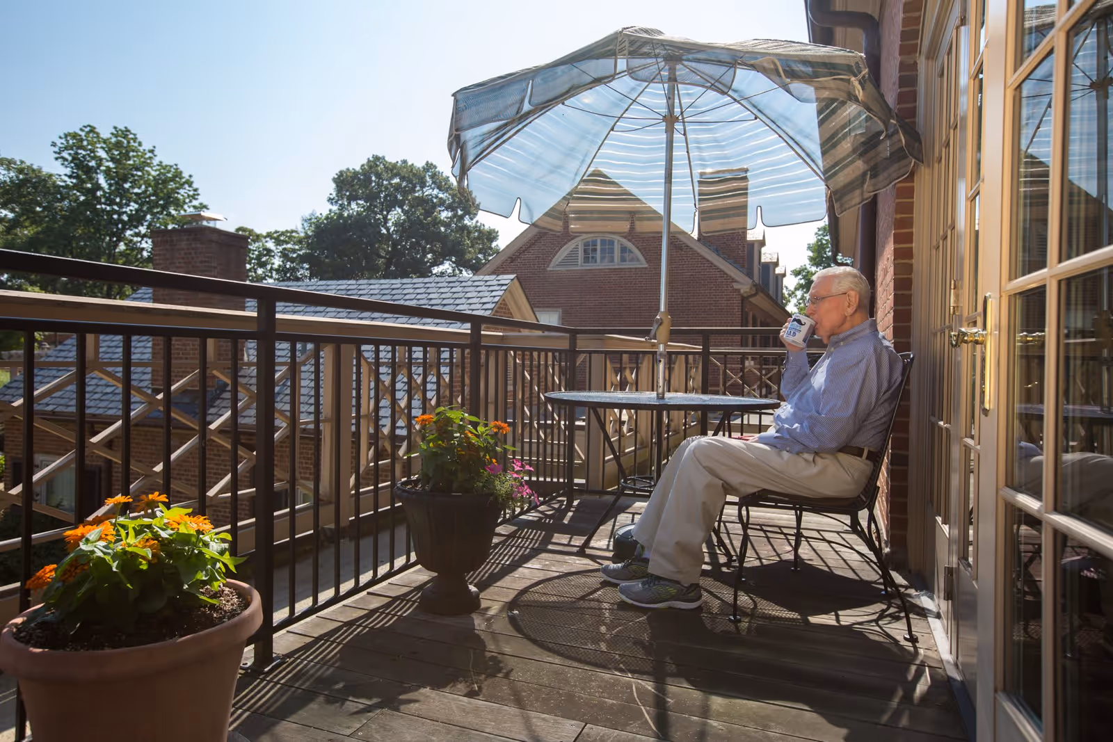 An older man sits on a sunlit balcony under a striped umbrella drinking from a mug, with potted flowers and nearby rooftops visible.