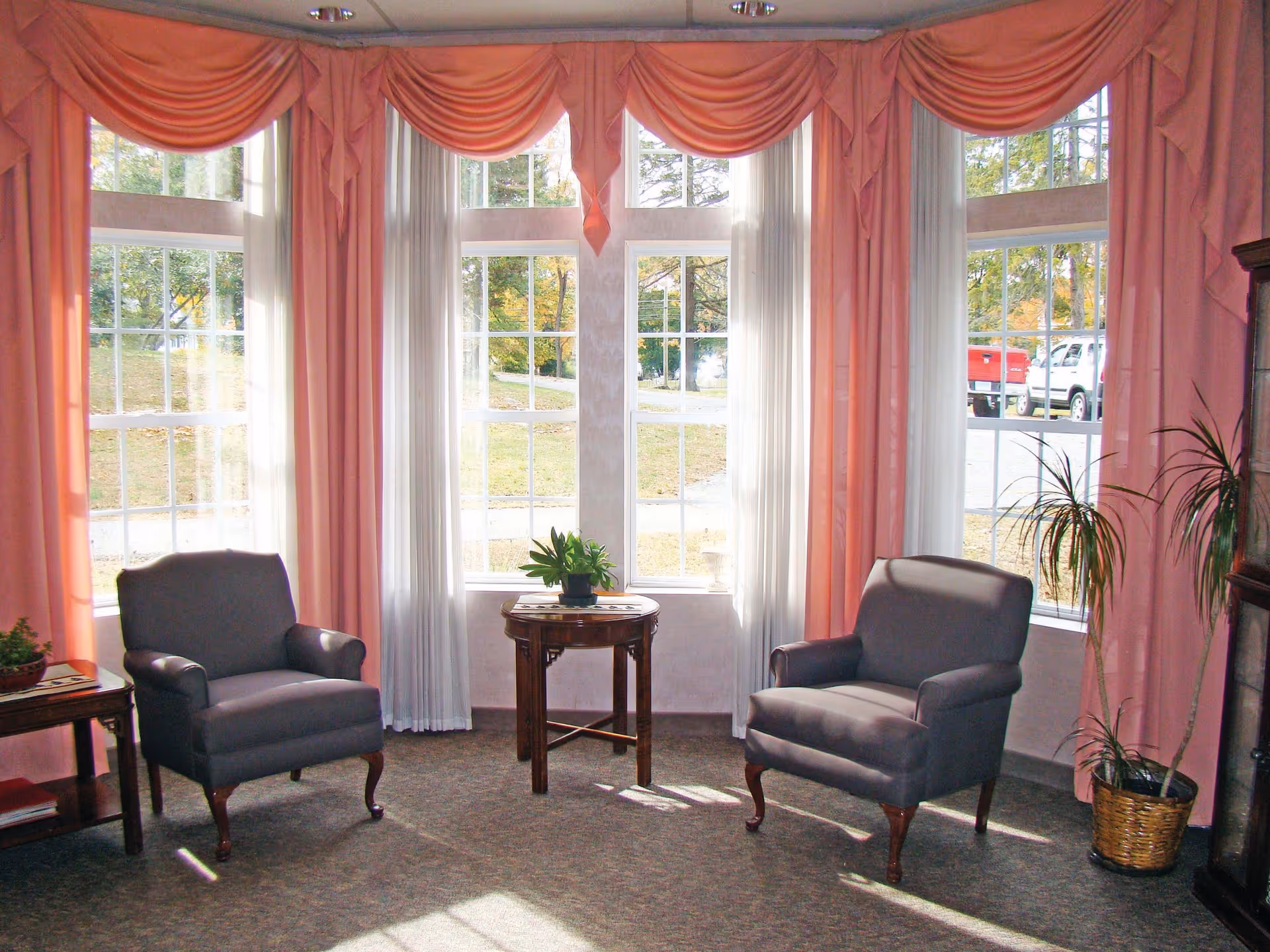 A cozy sitting area with two gray upholstered armchairs facing each other, separated by a small round wooden table with a potted plant on it. The room features large windows with white sheer curtains and pink drapes, allowing natural light to fill the space. There is a tall potted plant in a woven basket on the right side and a small wooden side table with another plant on the left side.