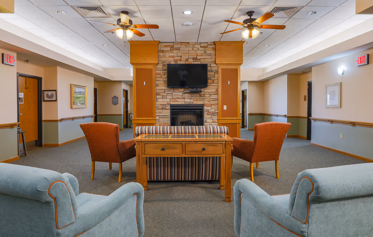 A cozy common area in an assisted living facility featuring a stone fireplace with a mounted flat-screen TV above it. The room has two ceiling fans with lights, two orange armchairs, a striped loveseat, a wooden table with drawers, and two light blue armchairs in the foreground. The walls are painted beige with a green lower section, and there are exit signs and framed artwork on the walls.