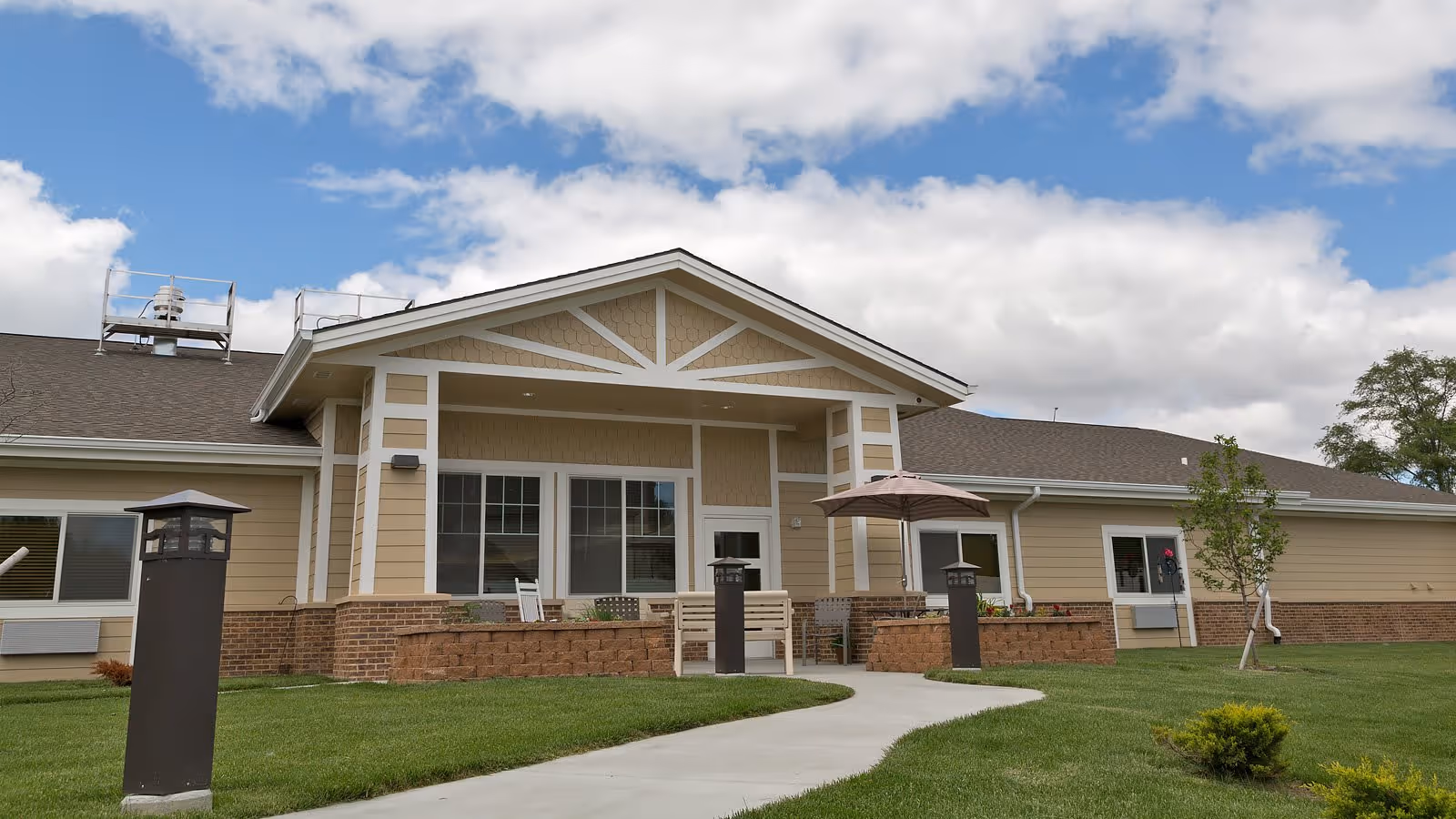 Exterior view of Hansen House, a single-story building with beige siding and brick accents. The building has a covered entrance with white trim and a peaked roof. There is a concrete walkway leading up to the entrance, surrounded by green grass and small shrubs. Outdoor seating with an umbrella is visible near the entrance under a partly cloudy sky.