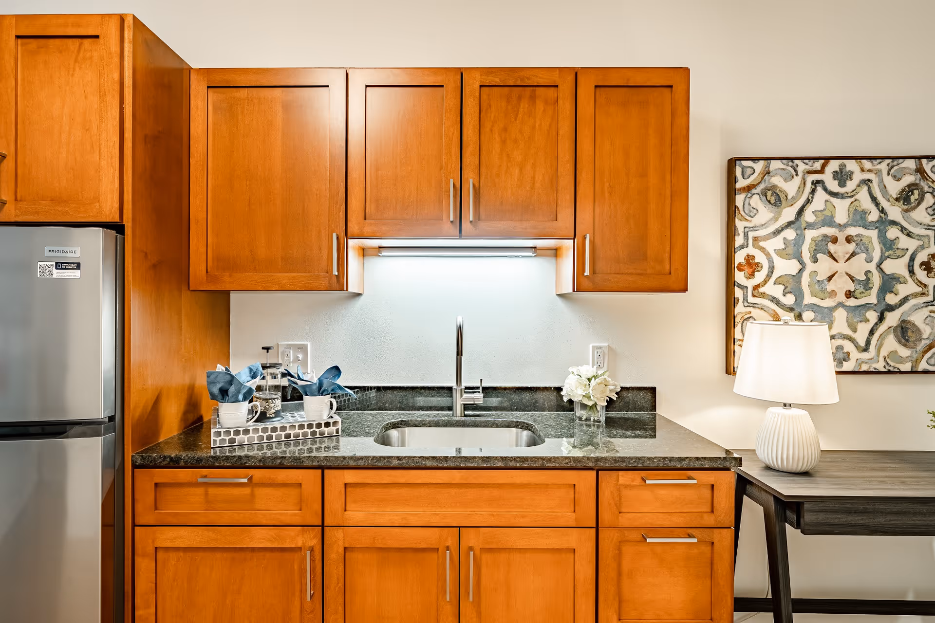A modern kitchen area with wooden cabinets, a stainless steel refrigerator, a granite countertop with a sink, a small tray holding cups and napkins, a vase with white flowers, a table lamp, and a decorative framed artwork on the wall.