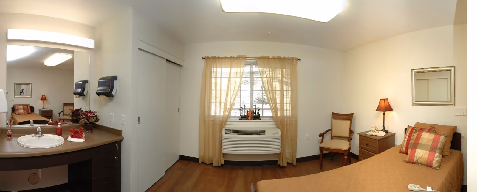 A senior living facility bedroom with a single bed covered in a brown quilt and a plaid pillow. Next to the bed is a wooden nightstand with a lamp and a chair beside it. A window with sheer beige curtains and an air conditioning unit is centered on the wall. On the left side, there is a sink with a large mirror above it and two wall-mounted soap or paper towel dispensers. The floor is wooden, and the walls are painted light beige.
