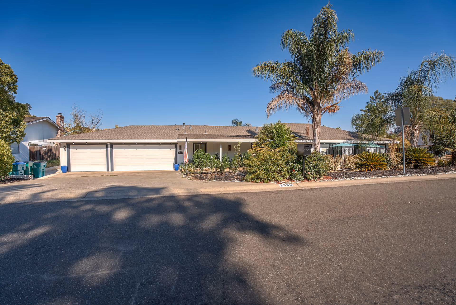 Single-story residential building with a three-car garage, surrounded by palm trees and other greenery under a clear blue sky.