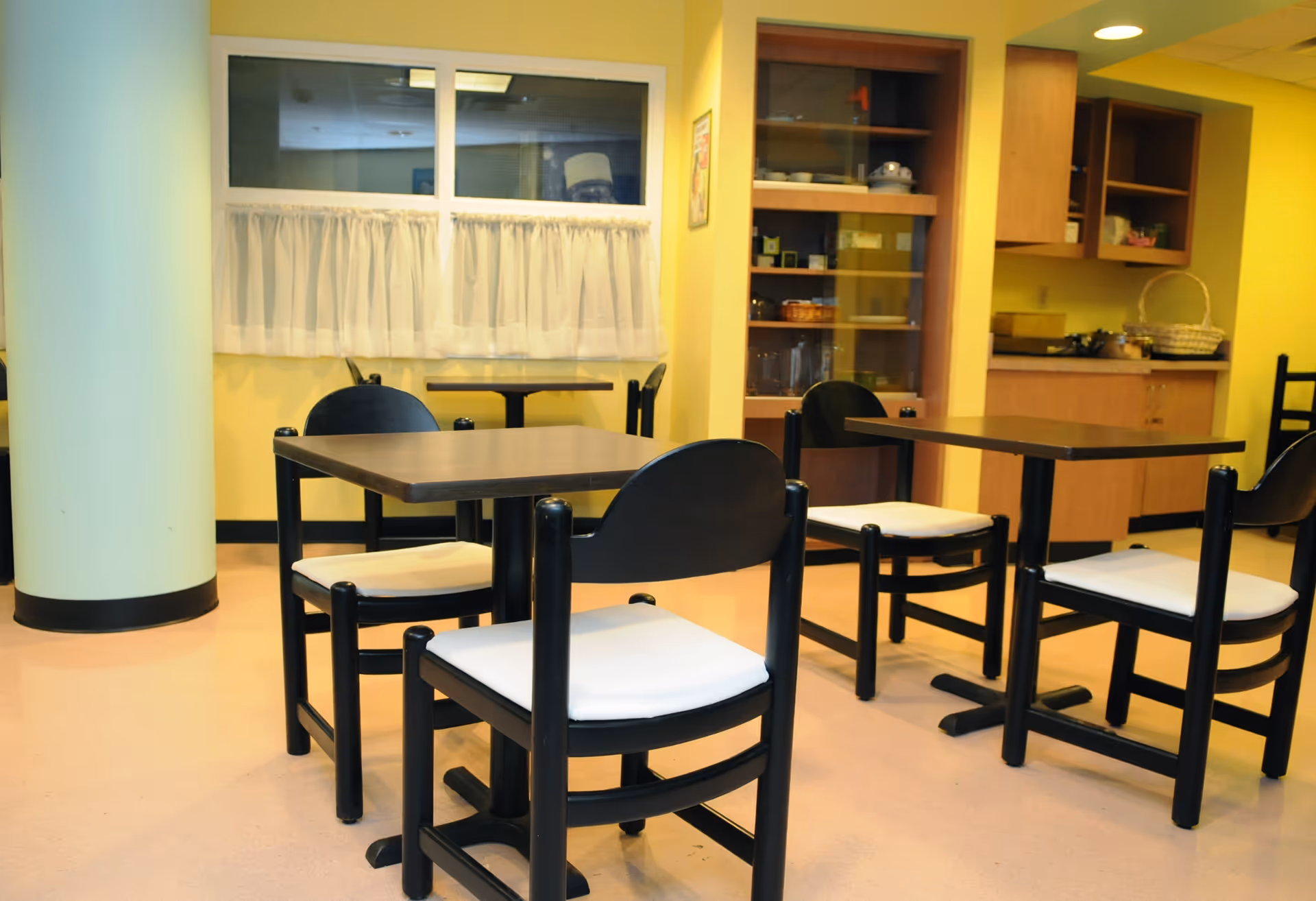 Interior view of a dining area with several black wooden tables and chairs with white cushions. The room has yellow walls, a large white column, a window with white curtains, and a wooden cabinet with glass doors displaying dishes and other items.