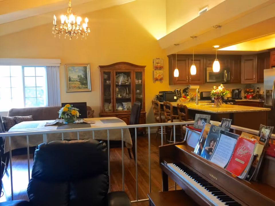 Interior view of a cozy living space featuring a piano with sheet music, a dining table with a flower centerpiece, a china cabinet, a kitchen island with hanging pendant lights, and a chandelier hanging from a vaulted ceiling.