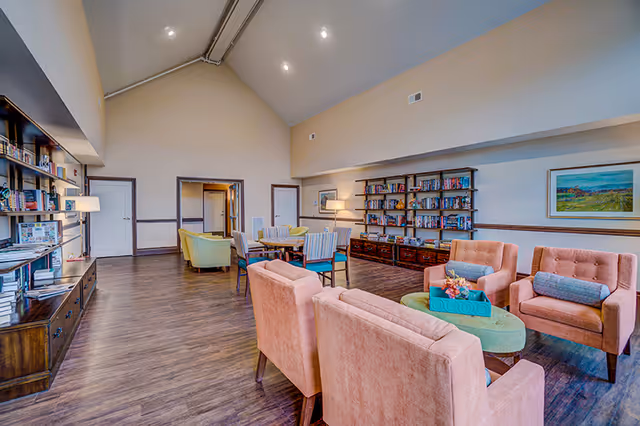 A spacious living room area in a senior living facility with high vaulted ceilings and wooden flooring. The room features multiple seating arrangements including pink upholstered armchairs around a green oval coffee table, a round table with chairs, and bookshelves filled with books along the walls. There is a framed landscape painting on the wall and soft lighting from lamps.