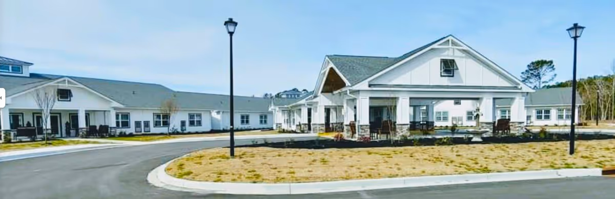Exterior view of a single-story senior living facility with white siding and gray roofs, featuring a covered entrance with seating areas and lamp posts along the driveway.