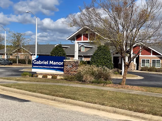 Exterior view of Gabriel Manor Assisted Living & Memory Care Neighborhood building with a large sign in front displaying the facility name. The building has a pitched roof, brick and siding exterior, and is surrounded by trees and landscaping under a partly cloudy sky.