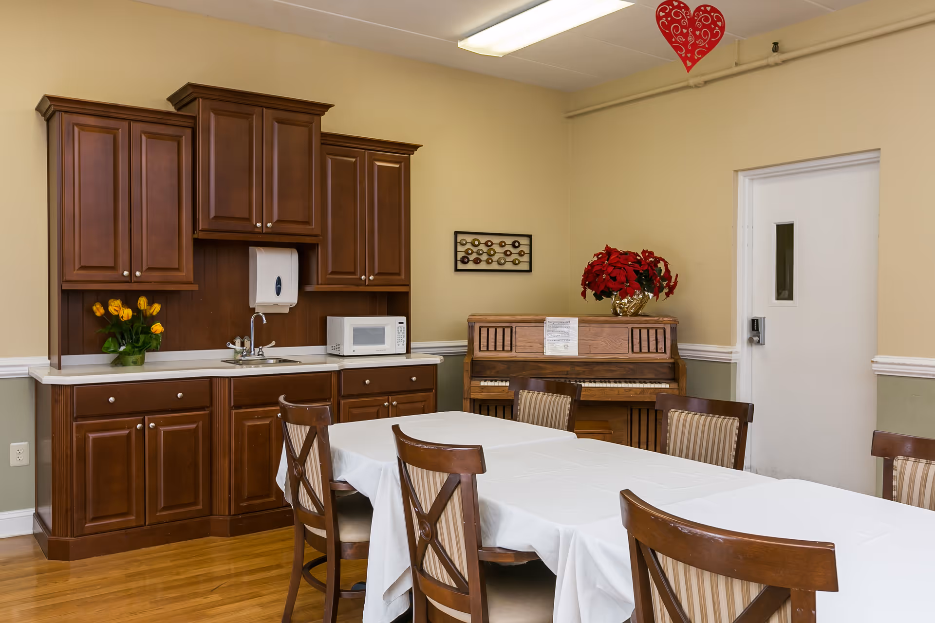 A dining area with a long table covered with a white tablecloth surrounded by wooden chairs with striped cushions. In the background, there is a wooden cabinet with a sink, a microwave, and a vase with yellow flowers. To the right, there is an upright piano with a red poinsettia plant on top and a white door with a small window. A red heart decoration hangs from the ceiling.