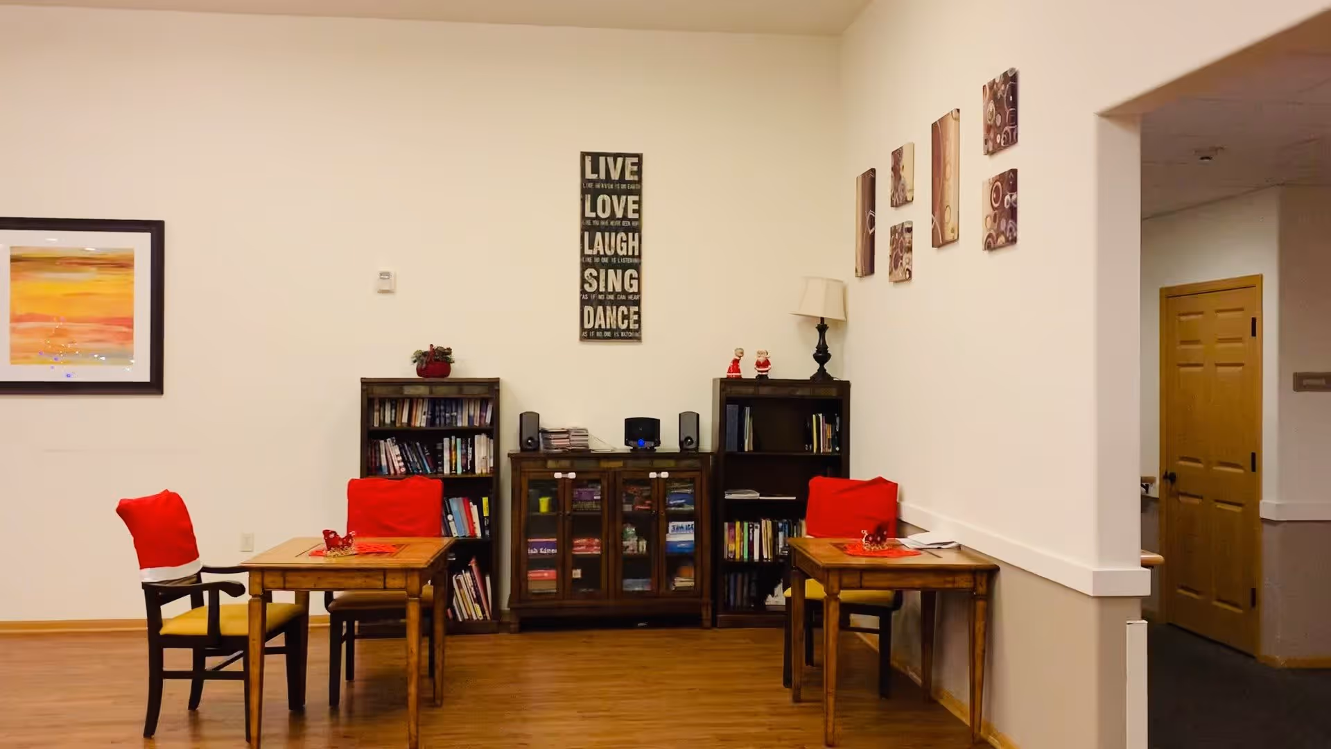 A cozy interior room with two wooden tables and chairs, each chair covered with a red cloth. There are bookshelves filled with books and decorative items against the wall, along with a cabinet in the center. Wall art includes a framed colorful painting, a vertical sign with inspirational words, and four smaller square pieces. The floor is wooden, and there is a doorway leading to another room.
