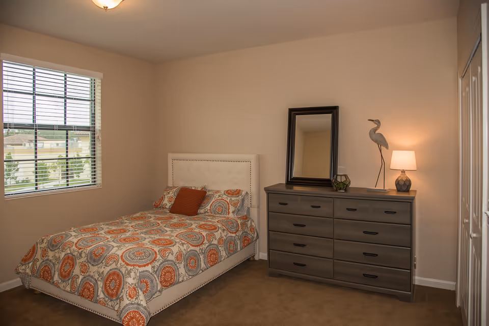 A bedroom with a bed covered in a patterned bedspread featuring orange and gray circular designs, a matching orange pillow, a window with blinds on the left wall, and a gray dresser with a mirror, a decorative bird sculpture, a small plant, and a lamp on top against the right wall.
