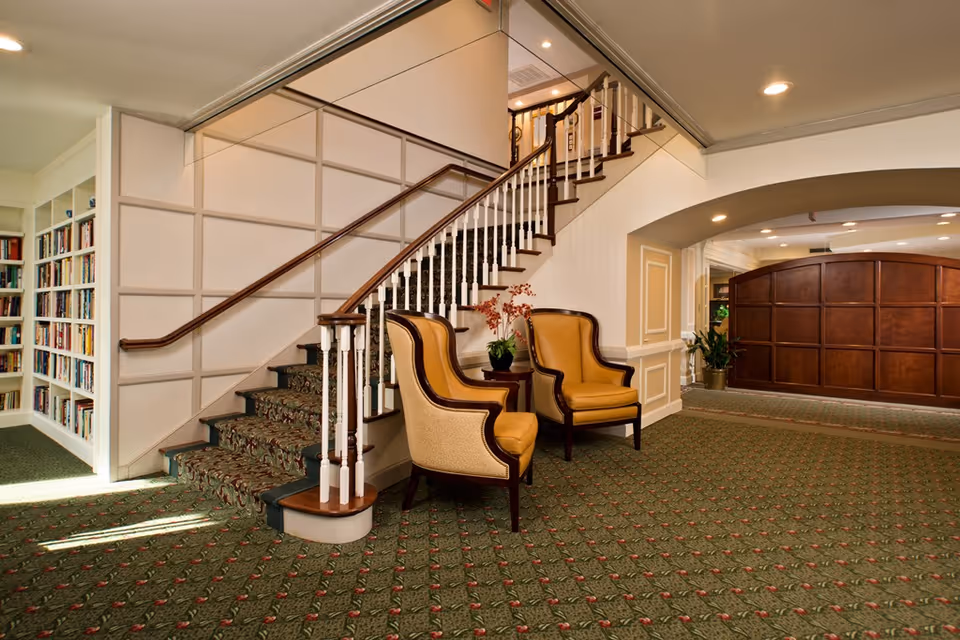 Carpeted lobby with a curved staircase, two upholstered armchairs, a bookshelf, and a wood-paneled reception desk.