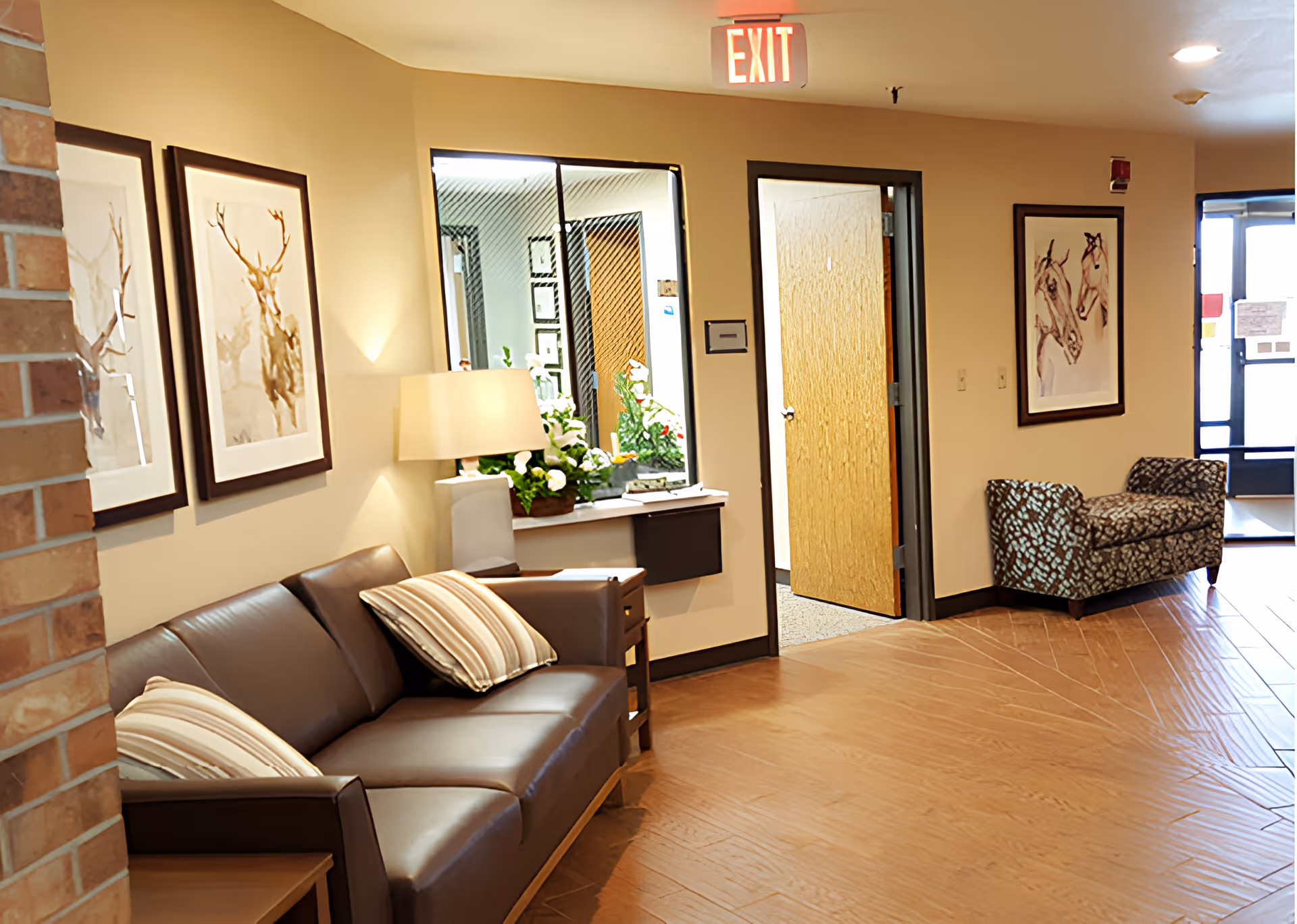 A cozy waiting area with a brown leather sofa adorned with striped pillows, a side table with a lamp and flowers, framed animal artwork on the walls, a patterned bench near the entrance door, and an exit sign above a doorway.