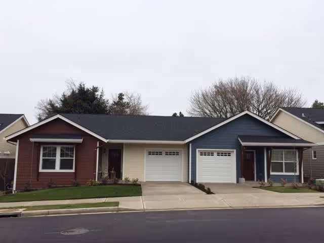 Exterior view of a single-story residential building with two attached units, each with a garage door and a small covered porch. The left unit is painted brown and beige, while the right unit is painted blue and beige. There are small lawns and sidewalks in front of the building, with trees visible in the background under an overcast sky.