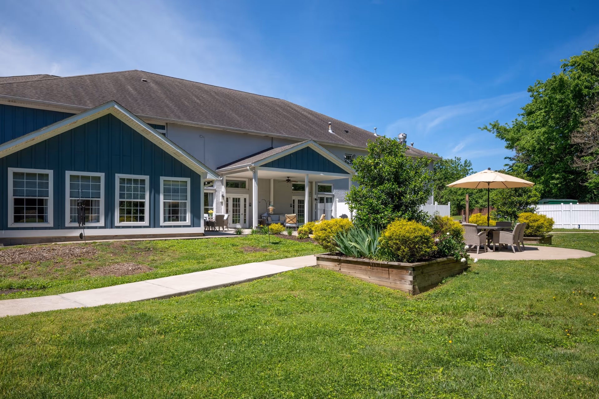Outdoor view of The Village at Bellevue assisted living facility showing a blue and white building with multiple windows, a covered patio area with chairs, a concrete pathway, green grass, a raised garden bed with bushes and plants, and a round table with chairs under a beige umbrella on a sunny day with a clear blue sky.