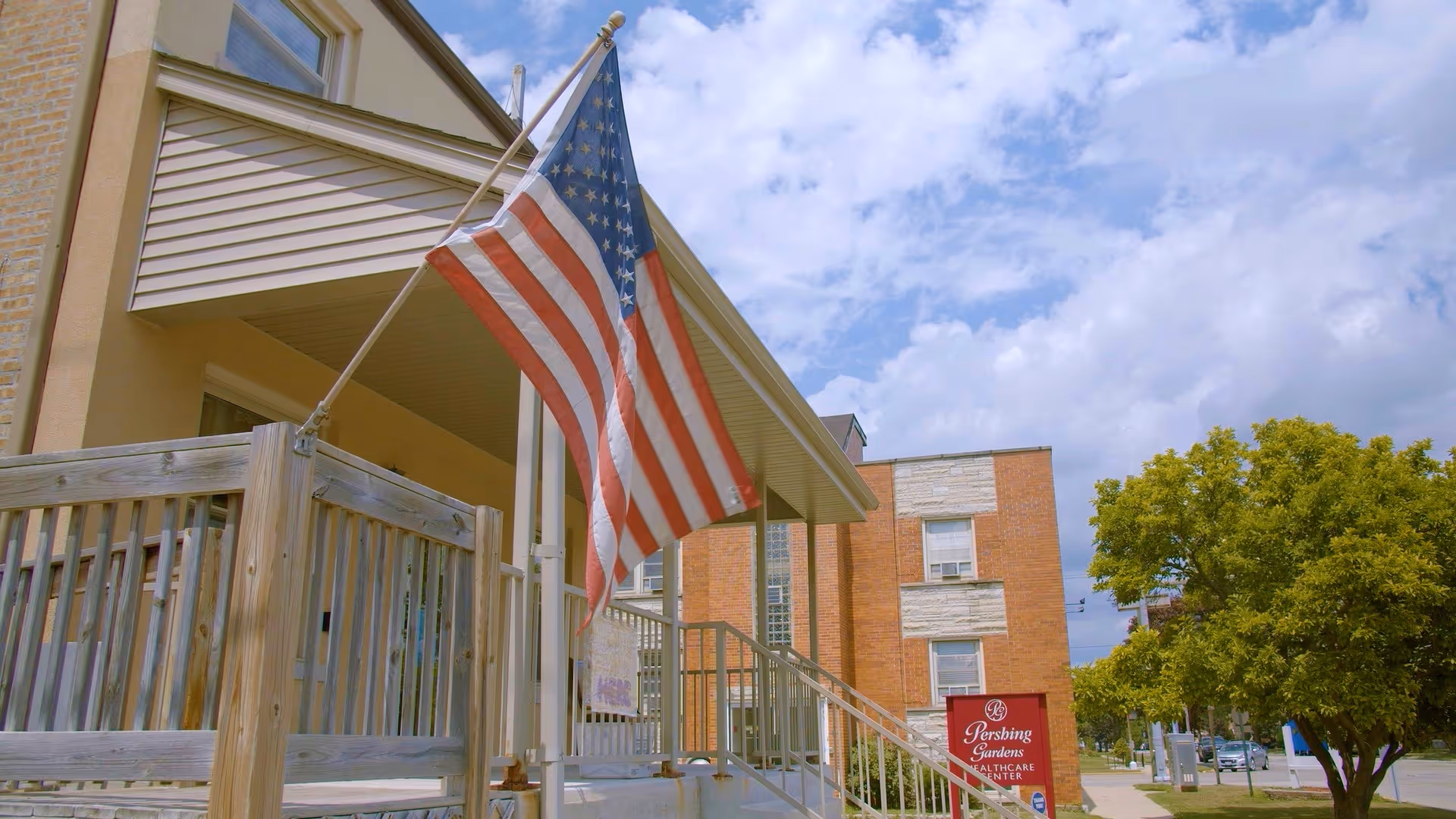 Exterior view of a senior living facility entrance with a wooden ramp and railing, an American flag mounted on a pole, a brick building in the background, a tree on the right side, and a red sign that reads 'Pershing Gardens Healthcare Center'. The sky is partly cloudy.