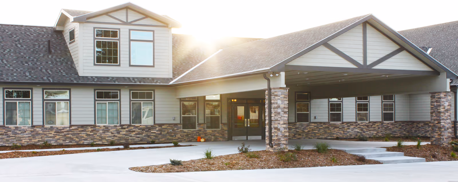 Exterior view of a modern single-story building with a covered entrance supported by stone pillars, multiple windows, and a gabled roof, with sunlight shining from behind.