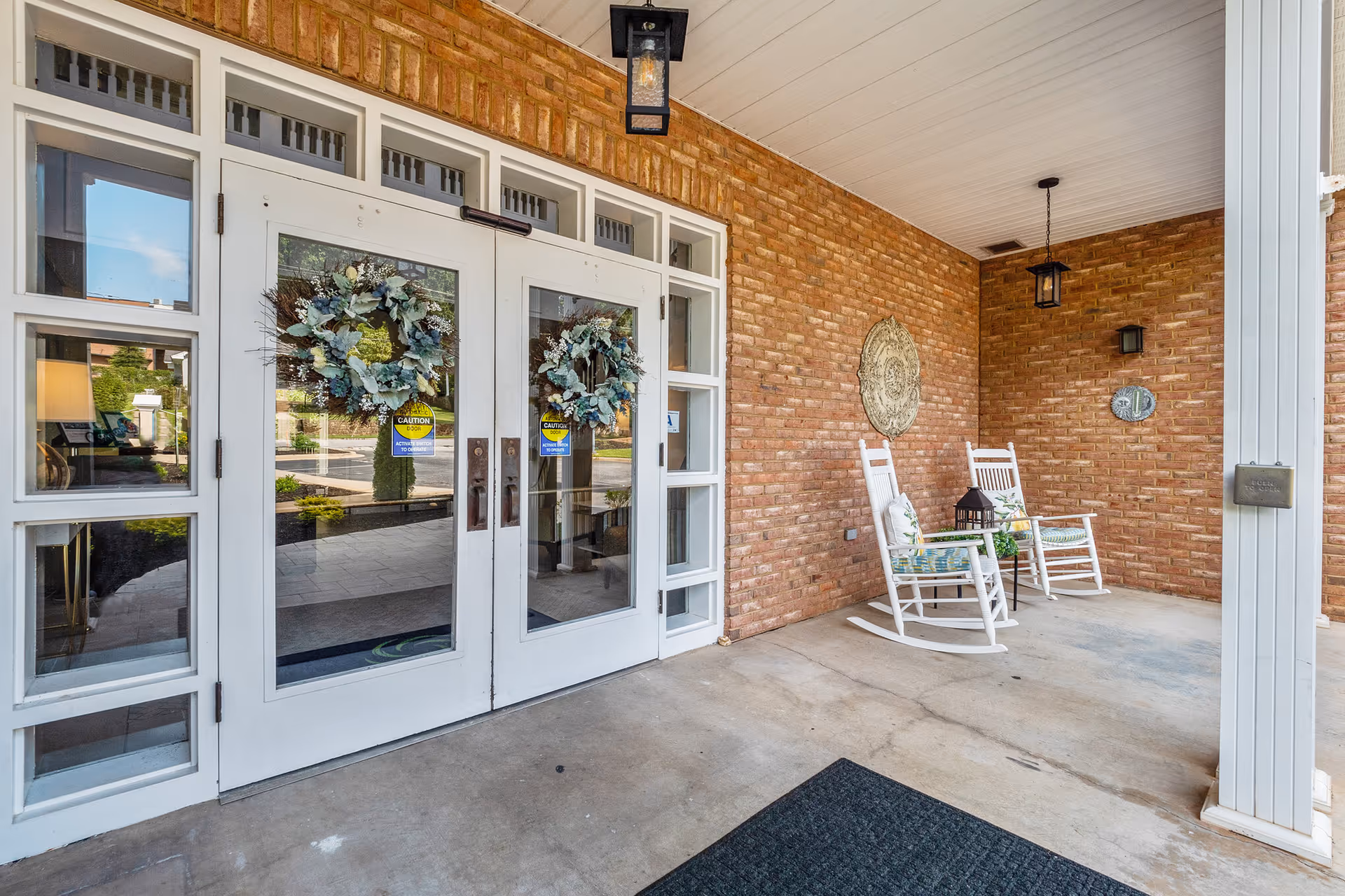 Covered entrance area of a building with double glass doors decorated with wreaths. Two white rocking chairs with cushions are placed against a brick wall, accompanied by a small table with a lantern. The area is well-lit with hanging and wall-mounted lantern-style lights.