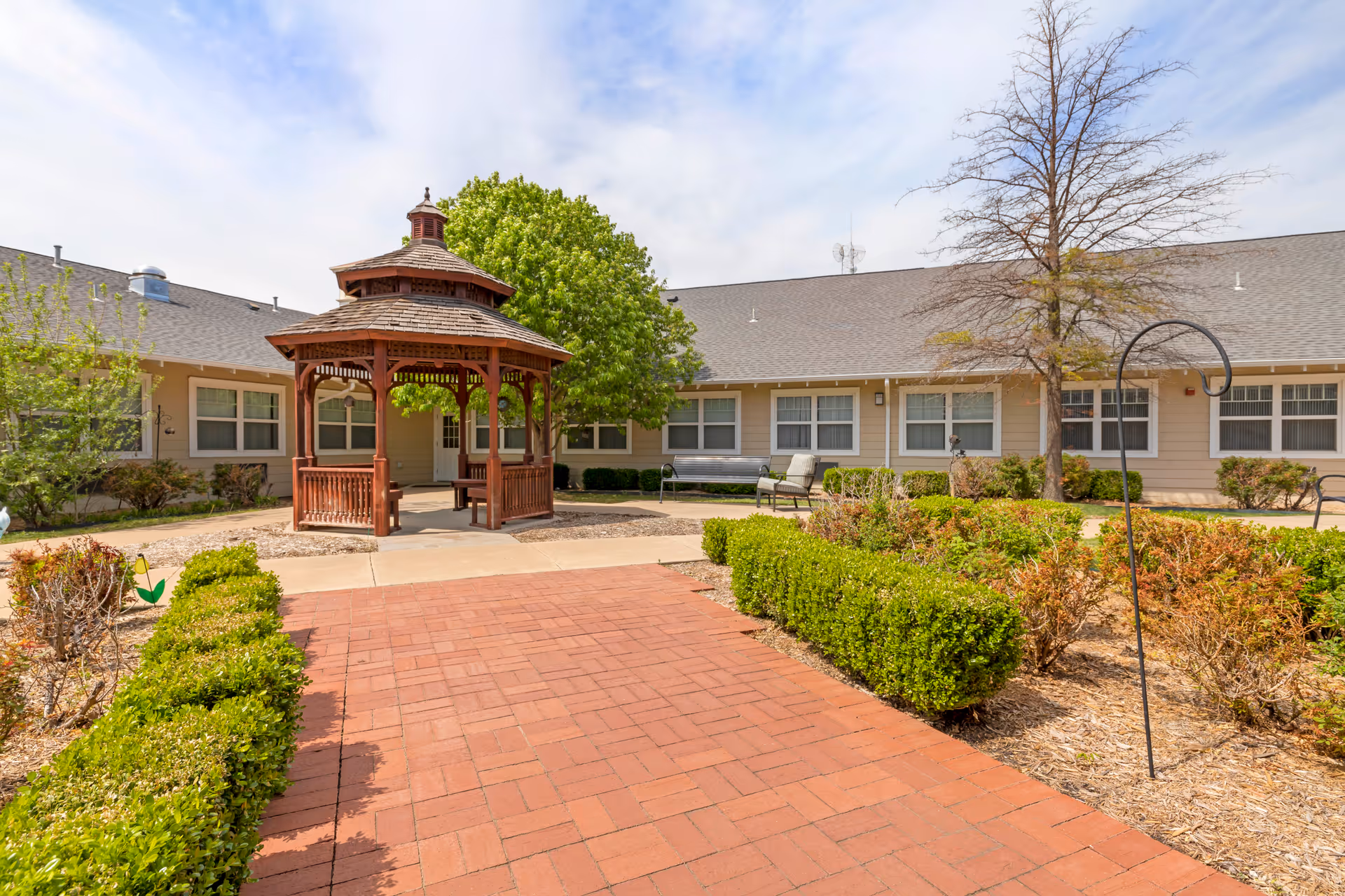 Outdoor courtyard area of Aberdeen Memory Care of Tulsa featuring a red brick pathway leading to a wooden gazebo surrounded by green bushes, trees, and benches with a beige building in the background under a partly cloudy sky.
