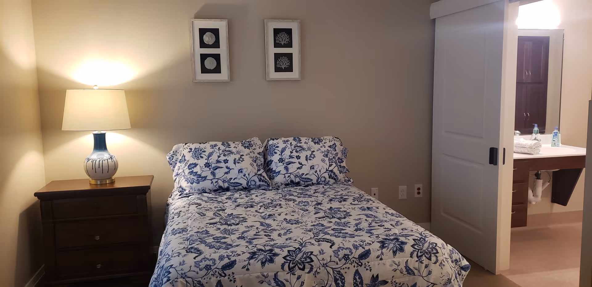 A bedroom with a bed covered in a blue and white floral patterned comforter and matching pillows. To the left of the bed is a wooden nightstand with a decorative lamp. Above the bed are two framed pictures with botanical designs. To the right, a sliding door is partially open, revealing a bathroom sink area with towels and toiletries.