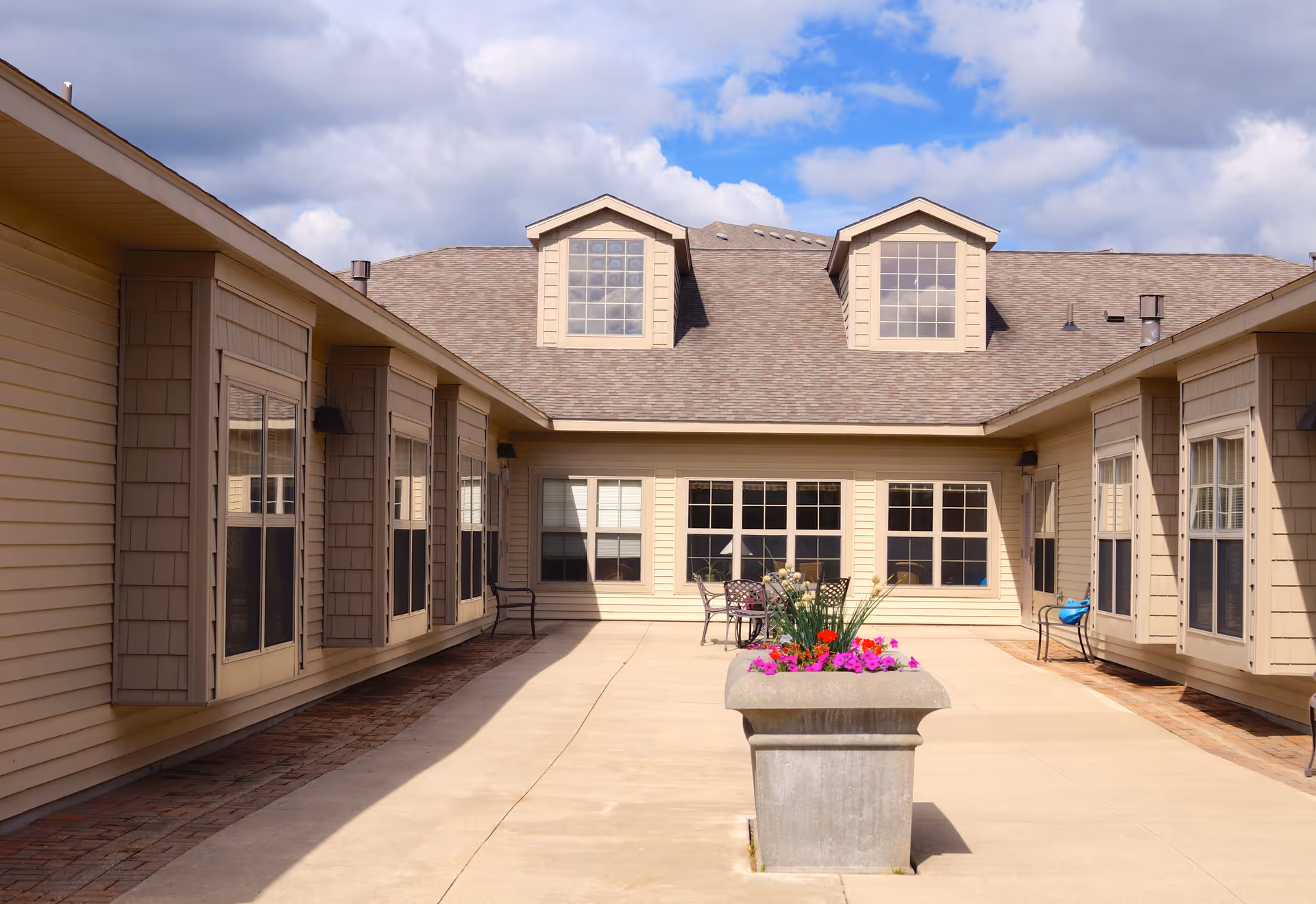 Outdoor courtyard area of Bay Ridge Assisted Living Center with beige siding buildings on both sides, large windows, a concrete walkway, a planter with colorful flowers in the center, and a table with chairs near the far building under a partly cloudy sky.