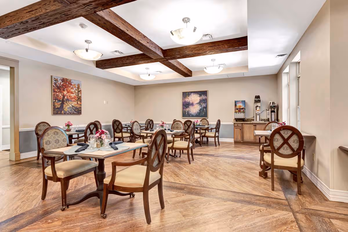 A bright dining room with wooden tables and upholstered chairs, exposed ceiling beams, wall art, and a beverage station.