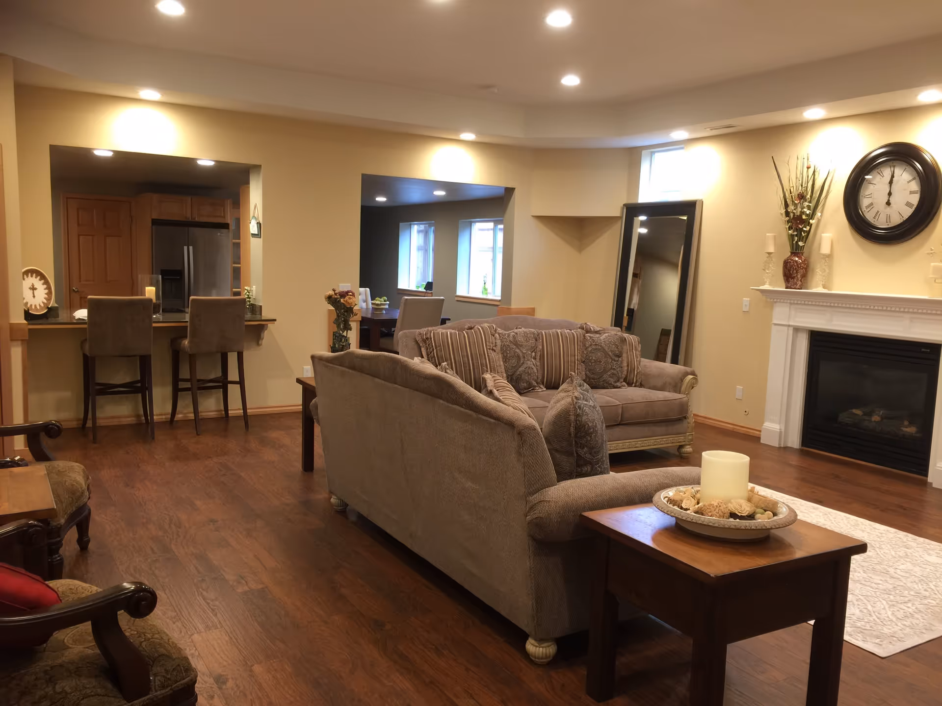 A cozy living room with two beige sofas adorned with patterned cushions, a wooden side table with a decorative candle centerpiece, a large wall clock above a white fireplace, and a tall mirror leaning against the wall. The room has wooden flooring and recessed ceiling lights. In the background, there is a kitchen area with a refrigerator and two bar stools at a counter, as well as a dining area with a table and chairs near windows.