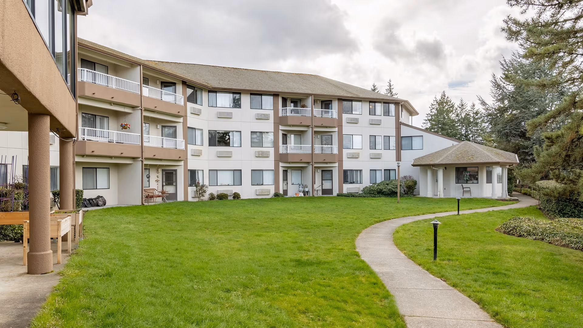 Exterior view of a multi-story retirement community building with balconies overlooking a well-maintained grassy courtyard and a curved concrete walkway leading to a small pavilion. Trees and shrubs surround the area under a cloudy sky.