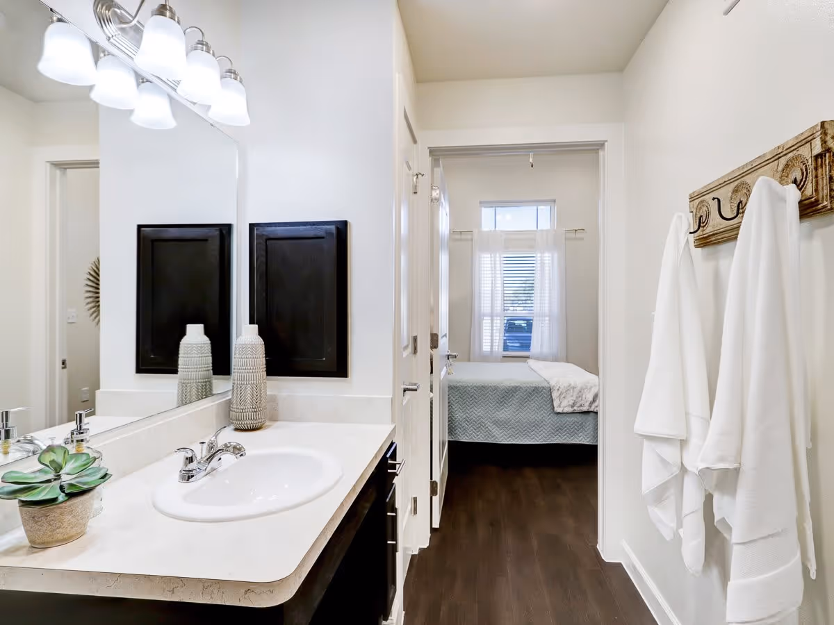 View of a bathroom with a white countertop, sink, and decorative items including a small plant and a vase. Above the sink is a large mirror with a light fixture featuring five lights. On the right wall, there is a wooden towel rack with three white towels hanging. Through an open door, a bedroom with a bed covered in a light blue quilt and a window with white curtains is visible.