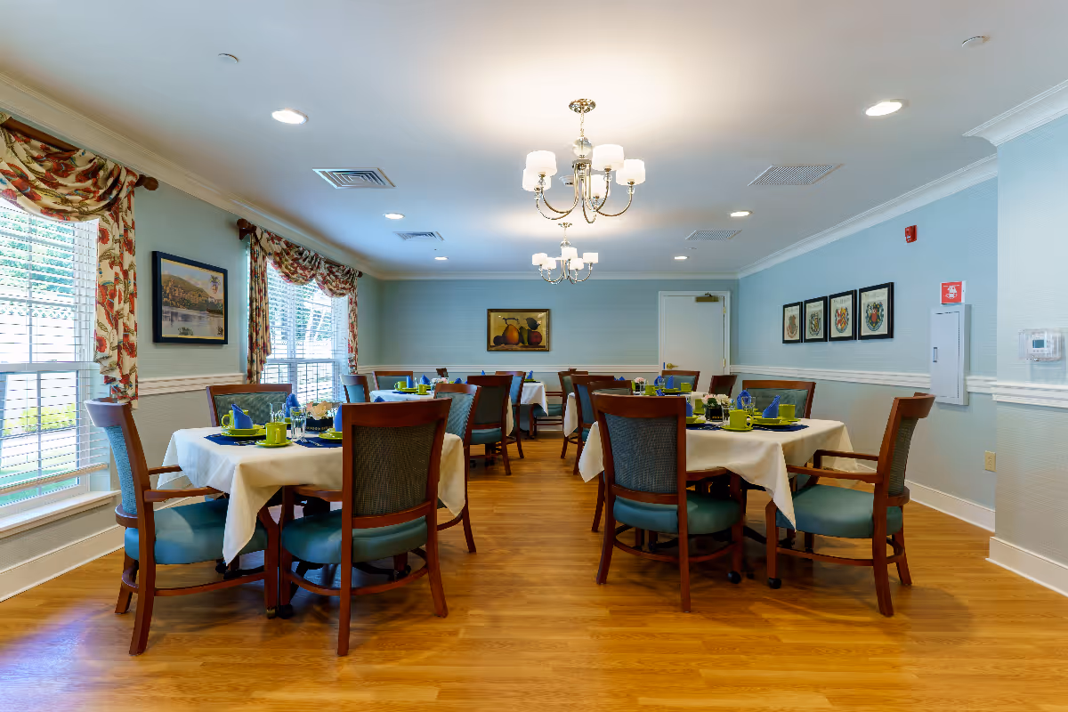 A bright dining room in a senior living facility with several tables covered in white tablecloths, each set with green cups, blue napkins, and plates. The room has wooden floors, light blue walls, floral curtains on the windows, and framed artwork on the walls. Two chandeliers hang from the ceiling providing warm lighting.