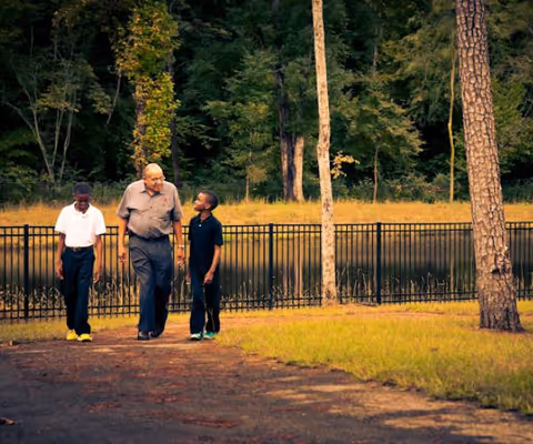 An elderly man walking outdoors on a paved path with two young boys, surrounded by trees and a black metal fence near a body of water.