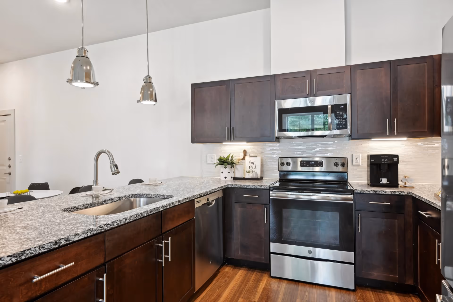 Modern kitchen with dark wood cabinets, stainless steel appliances including a stove, microwave, dishwasher, and refrigerator. Granite countertops with a built-in sink and two hanging pendant lights above the counter. A small plant and a coffee maker are on the countertop.