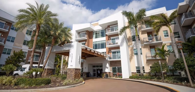 Exterior view of a multi-story senior living facility with balconies, palm trees, and a covered entrance under a partly cloudy sky.
