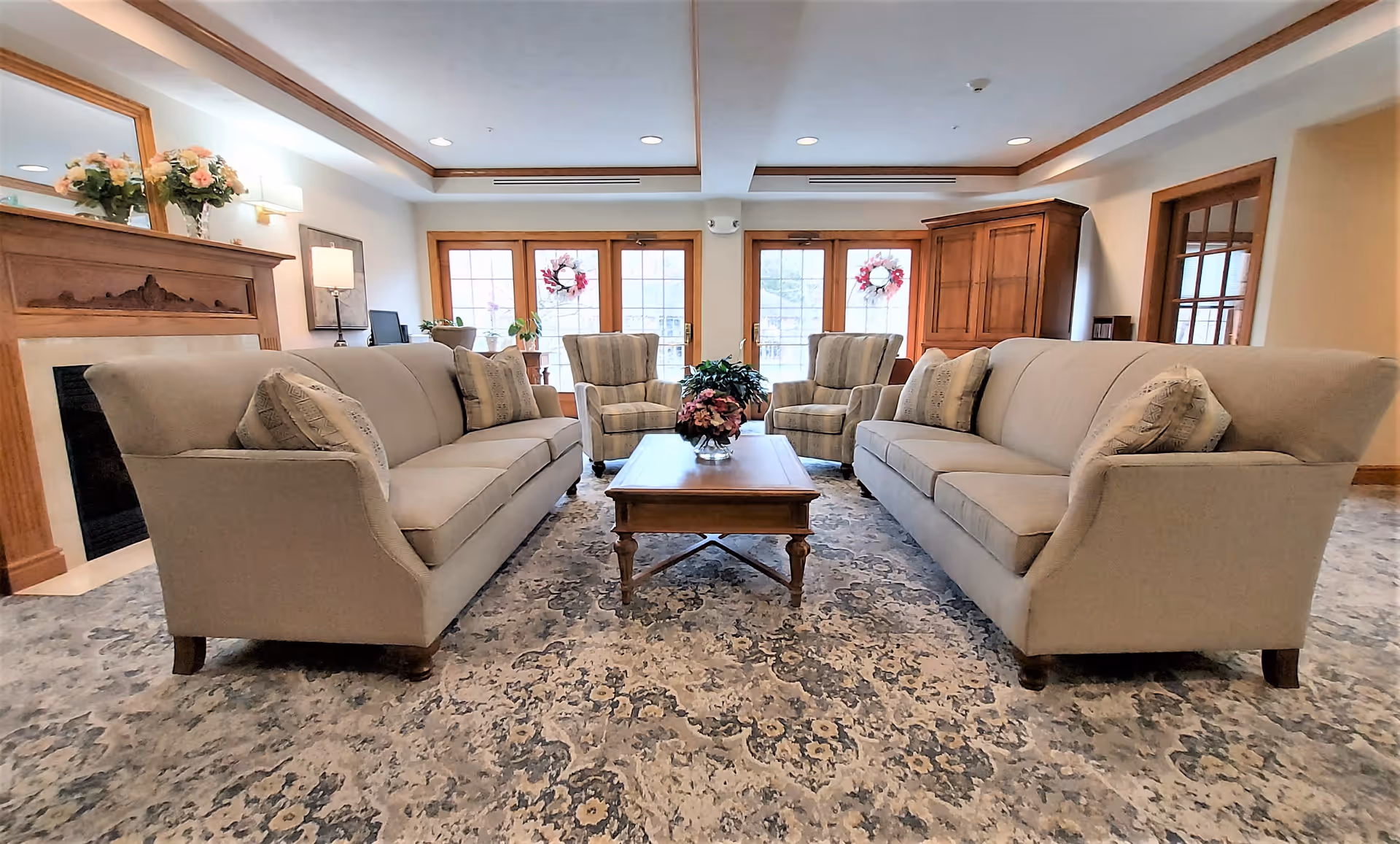A cozy living room with two beige sofas facing each other, a wooden coffee table with a flower arrangement in the center, two striped armchairs in the background, a fireplace with a mirror and flowers on the mantel, and large windows with wooden frames letting in natural light.