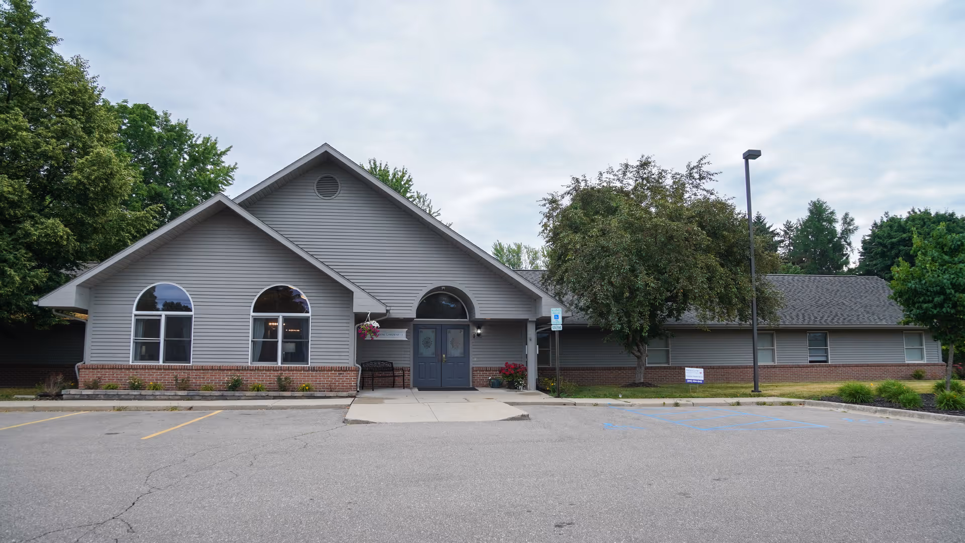 Exterior view of a single-story senior living facility building with gray siding and brick accents. The building has a peaked roof, arched windows, a double-door entrance, and a small bench near the entrance. There are trees and landscaping around the building, and an empty parking lot in front.
