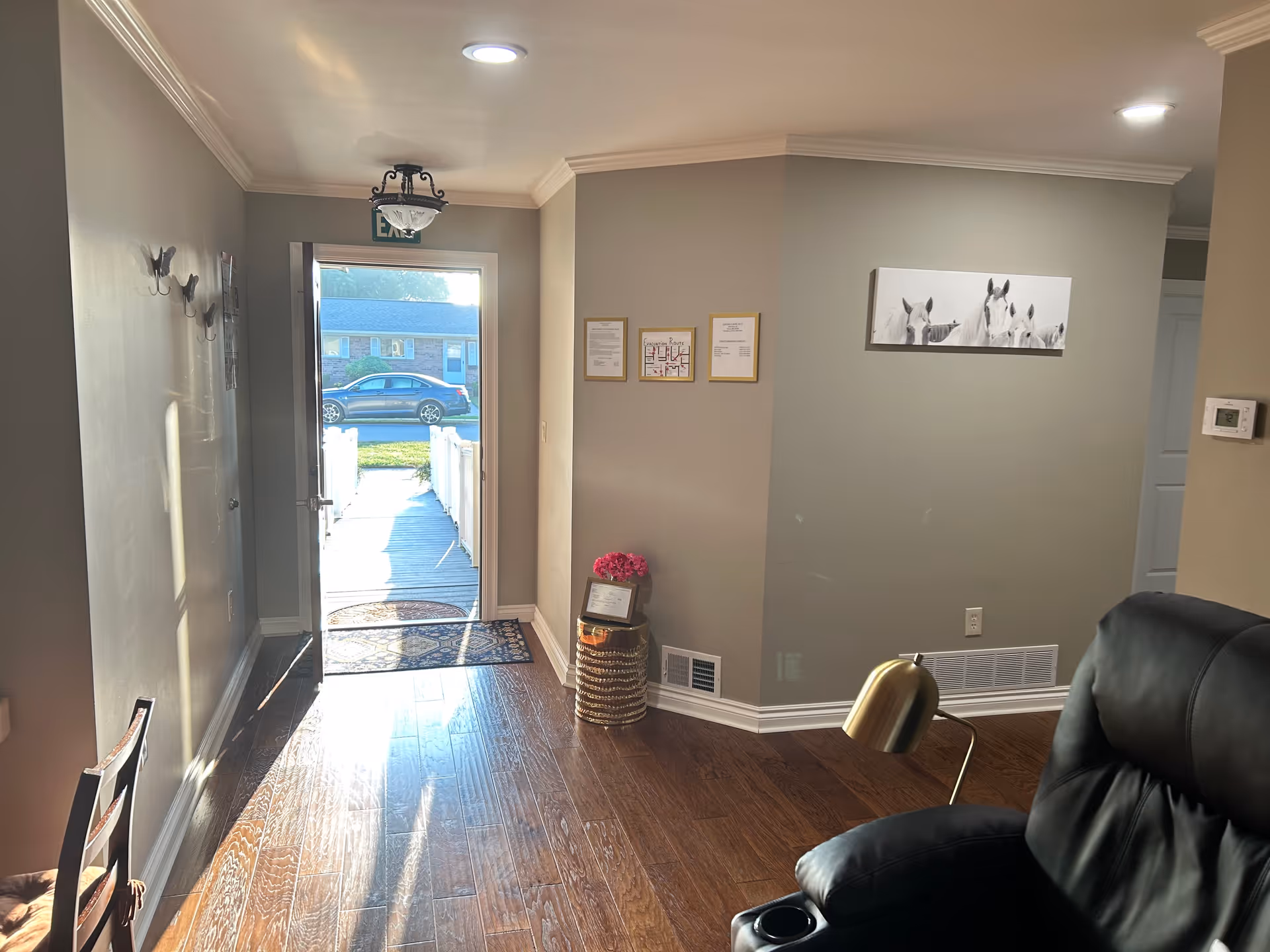 Interior view of a living area in a senior care facility with wooden flooring, a black leather recliner chair with a brass reading lamp, a small table with a flower pot and framed documents on the wall. The front door is open, showing a walkway outside with a parked car and a house across the street. The walls are painted light gray and decorated with a black and white horse painting.