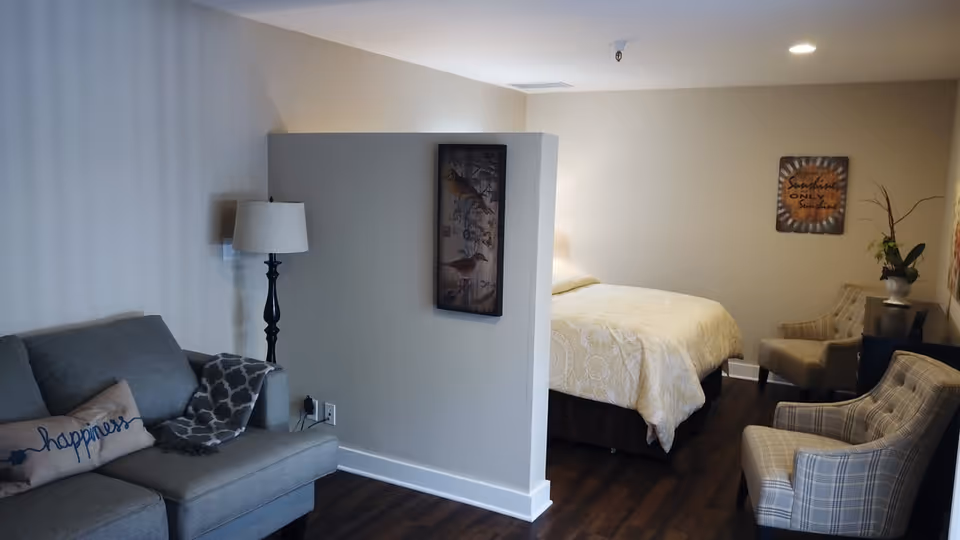 A cozy senior living room and bedroom area separated by a half wall. The living room has a gray couch with a pillow that says 'happiness', a floor lamp, and a patterned throw blanket. The bedroom features a bed with a light-colored comforter, two plaid armchairs, a small table with a plant, and wall art including a sign that reads 'Sunshine only sunshine'. The flooring is dark wood and the walls are light beige.