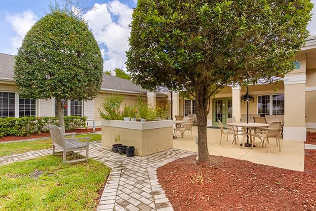 Outdoor patio area at a senior living facility with neatly trimmed trees, a wooden bench, potted plants, and tables with chairs under a covered porch. The building exterior is light-colored with large windows and a tiled walkway.