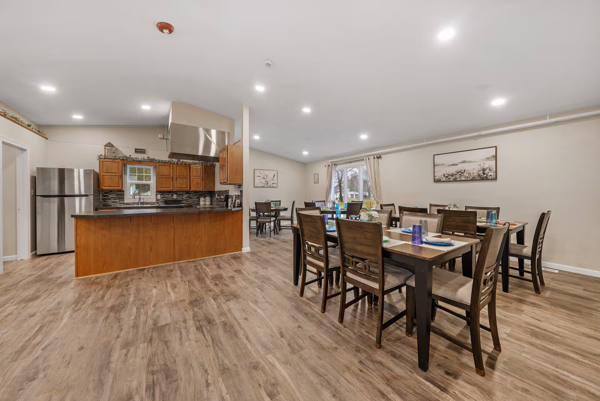 A spacious dining area with several wooden tables and chairs set with placemats, plates, and blue drinking glasses. The room features wood-look flooring, neutral-colored walls, and a large window with curtains letting in natural light. To the left, there is an open kitchen with wooden cabinets, a stainless steel refrigerator, a stove with a large hood, and a countertop bar area.