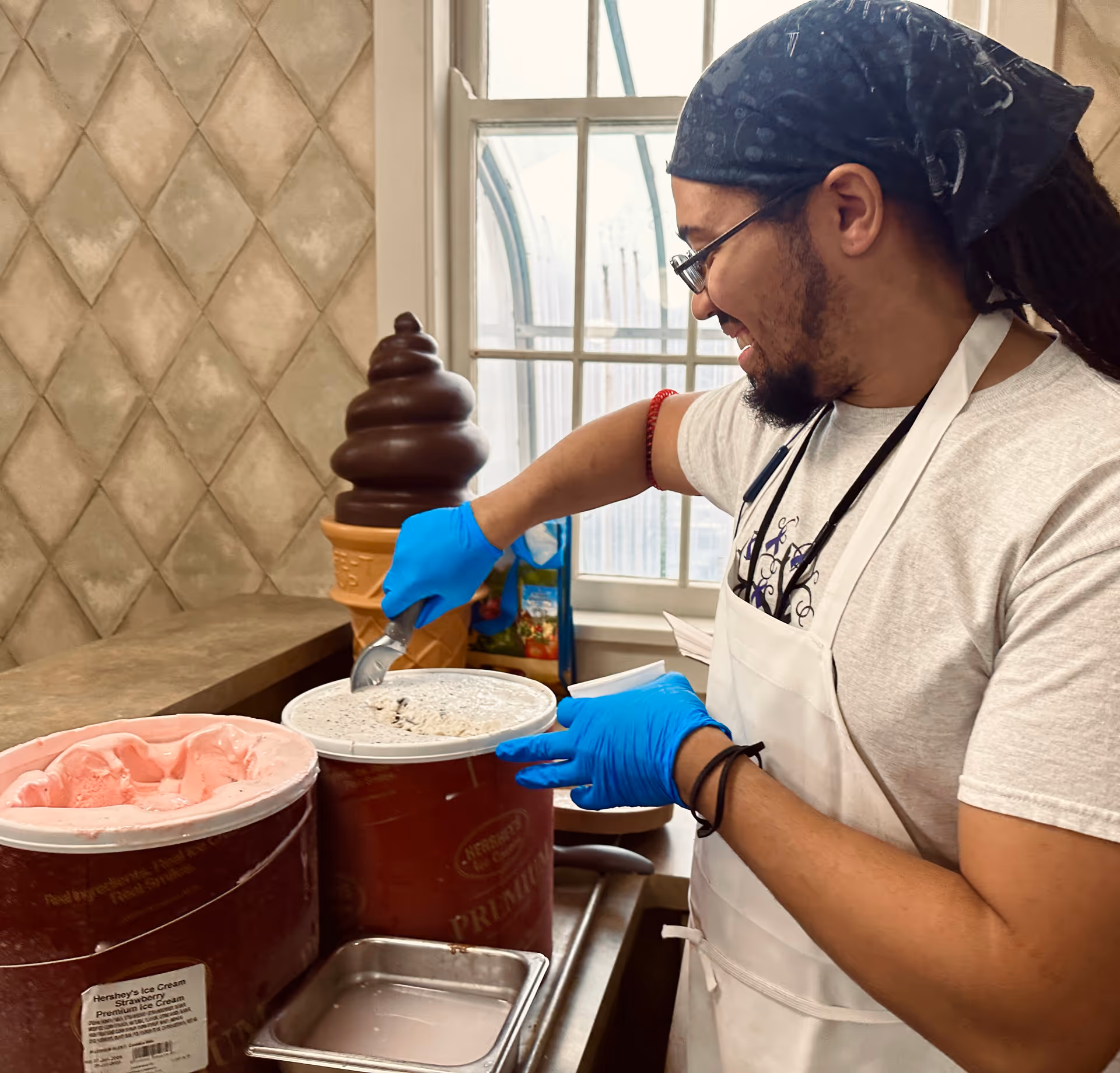 A man wearing a white apron, blue gloves, glasses, and a blue bandana is scooping cookies and cream ice cream from a large container. Next to it is a container of strawberry ice cream. Behind him is a window and a large decorative chocolate ice cream cone.