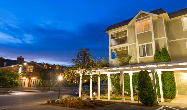 Exterior view of La Conner Retirement Inn at dusk, showing a multi-story building with balconies, a pergola-covered walkway, trees, and street lamps illuminating the area.