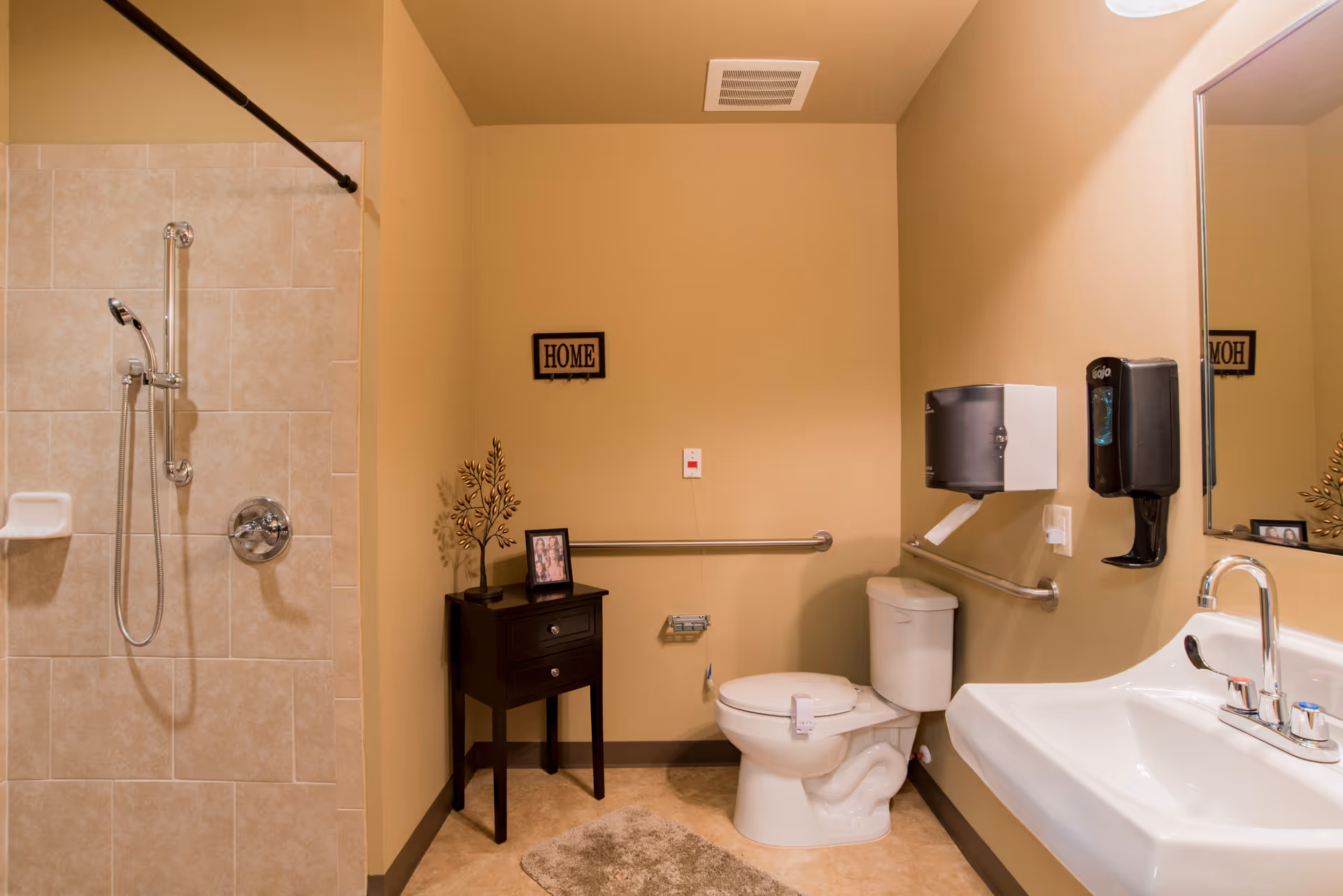 Accessible bathroom with a walk-in tiled shower, toilet with grab bars, sink, and a small side table under a 'HOME' sign.