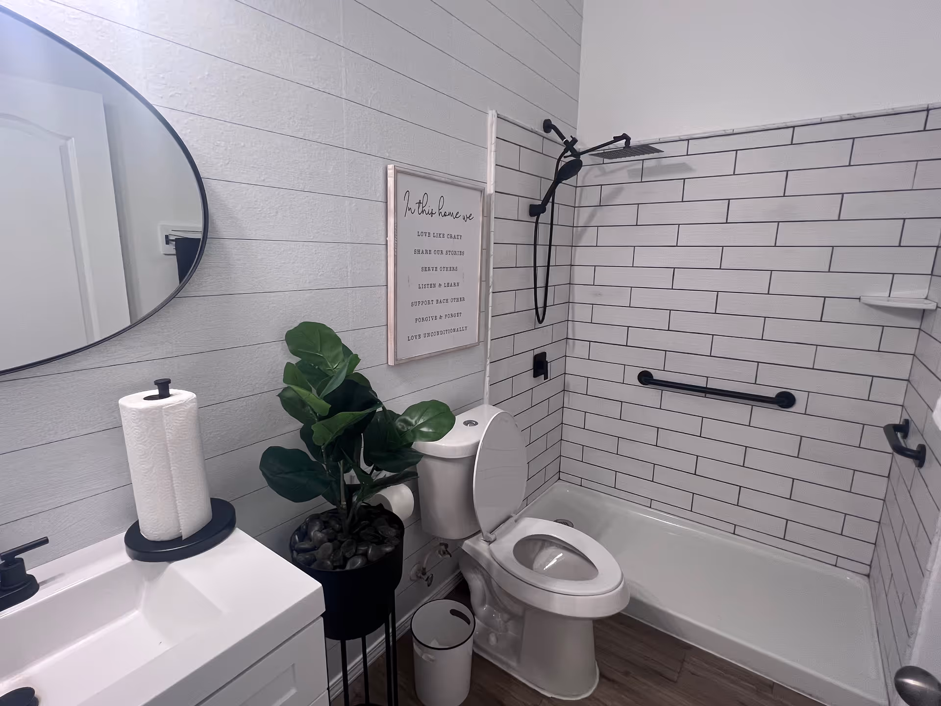 Modern bathroom with white tiled shower area featuring a black showerhead and grab bar. There is a white toilet with the lid open next to a small white sink with a paper towel holder. A green potted plant sits between the toilet and sink. A round mirror hangs on the wall above the sink, and a framed inspirational quote is mounted above the toilet.