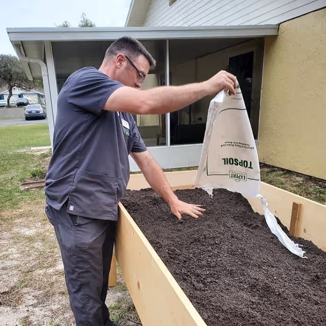 A man wearing dark scrubs is outdoors, pouring soil from a bag labeled 'Topsoil' into a raised wooden garden bed. He is spreading the soil with one hand while holding the bag with the other. The background shows part of a building with windows and a grassy area.