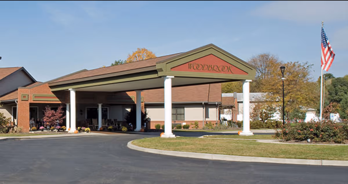 Exterior view of Woodbrook Assisted Living Residence, Inc. showing the main entrance with a covered driveway supported by white columns. The building is brick with a brown roof, and there is an American flag on a flagpole to the right. Landscaping with bushes and trees is visible around the entrance.