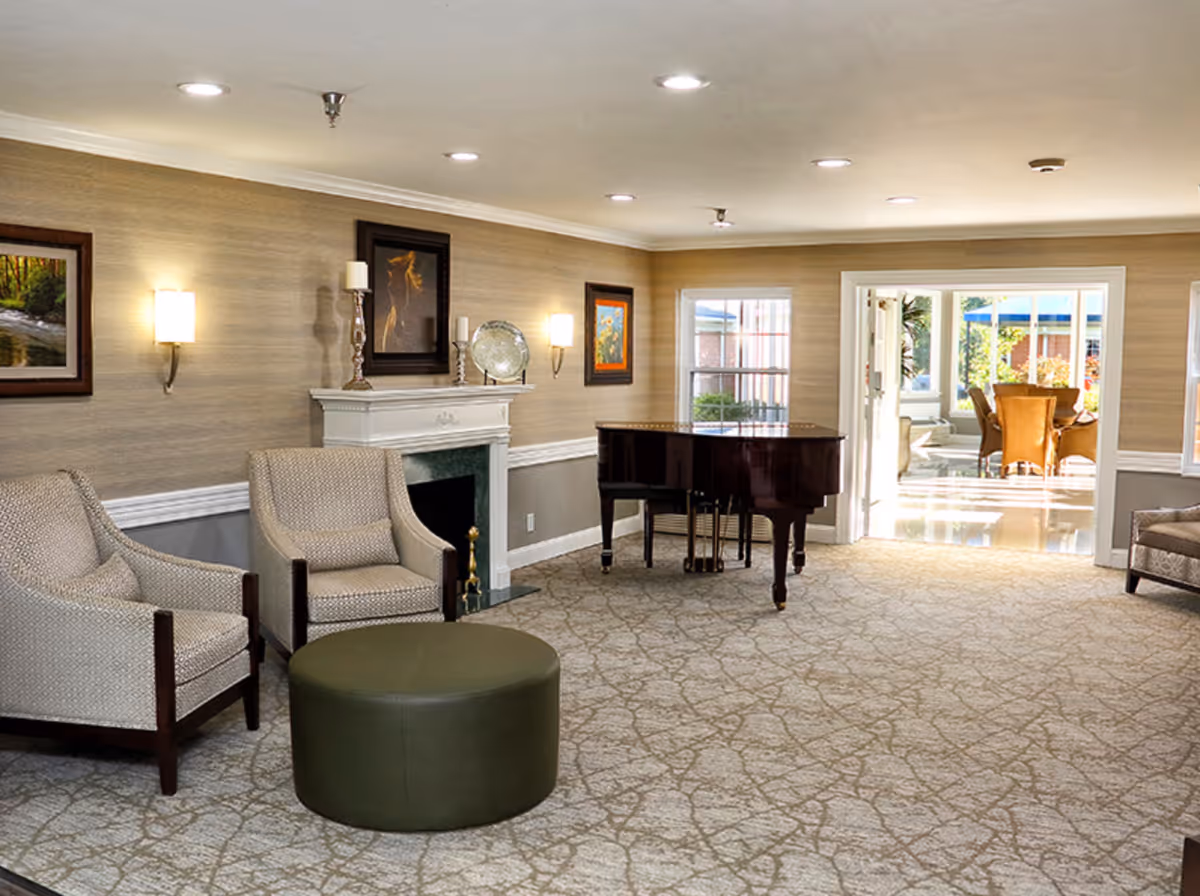 Bright communal lounge with armchairs, a round ottoman, fireplace, and a grand piano opening to a sunlit dining area.